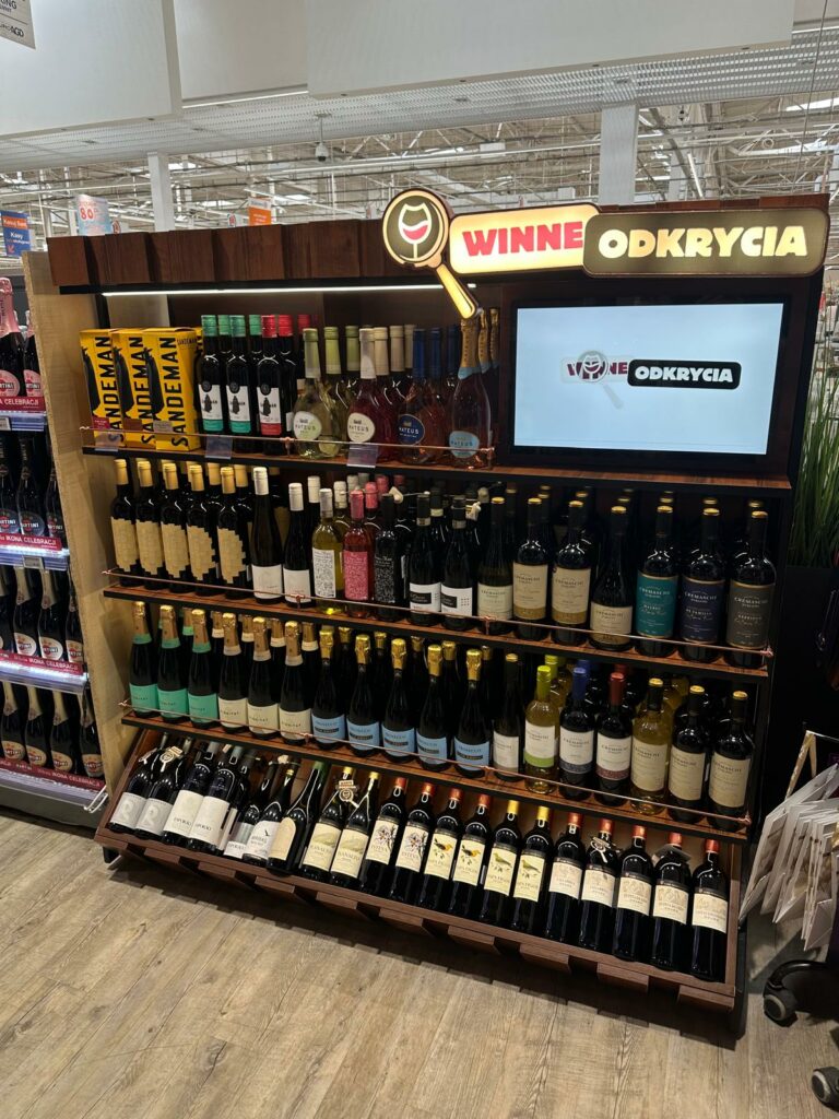 Permanent wine display with integrated digital screen, LED lighting and wooden shelving for retail environments.